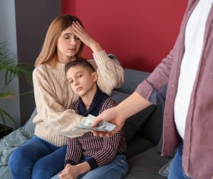 Stressed mother holds a child on the couch as a man offers cash support—breakup trauma isn't healing and the family tension is still heavy