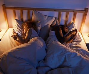 Couple lying back to back in bed on their phones at night, showing loss of sexual desire in marriage and why it matters to learn how to talk about sex with your partner.